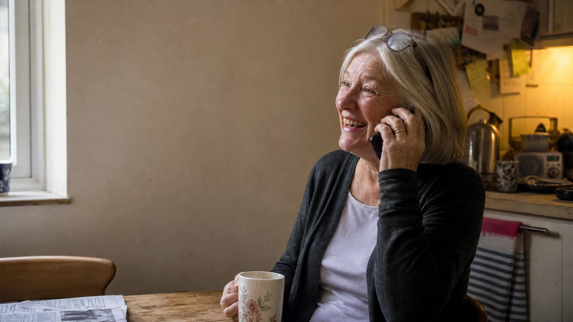 A woman in her late sixties laughing on the phone at her kitchen table